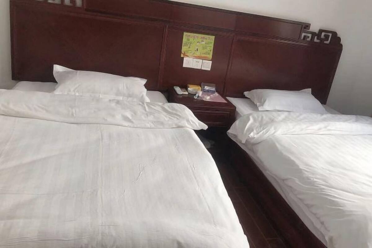 Habitación de hotel con dos camas individuales, cabecero de madera, mesita de noche con un teléfono y un objeto peque ño, y un anuncio colgado en la pared.