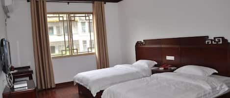 A hotel room with two single beds, a wooden headboard, a desk with a chair, and a window with curtains.