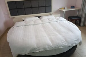 Uma cama bem arrumada com cabeceira, uma mesinha com uma cadeira e uma janela com cortinas.