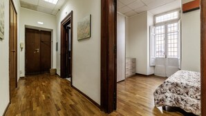 A hotel room with a wooden floor, a bed with a floral bedspread, a window with a view, a door, and a wall-mounted painting.