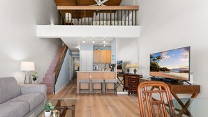 A spacious living area with a sofa, a glass coffee table, and a wooden chair. A kitchen with a bar area is visible in the background. - Convenient to downtown Kihei, across famous Charley Young Beach, 3 pool, tennis courts (MV3407) (Kihei)