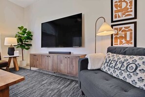 Una sala moderna con un televisor de pantalla plana en la pared, un sofá gris con un cojín que dice 'HELLO', un mueble de madera, una mesita con una lámpara y una planta verde.
