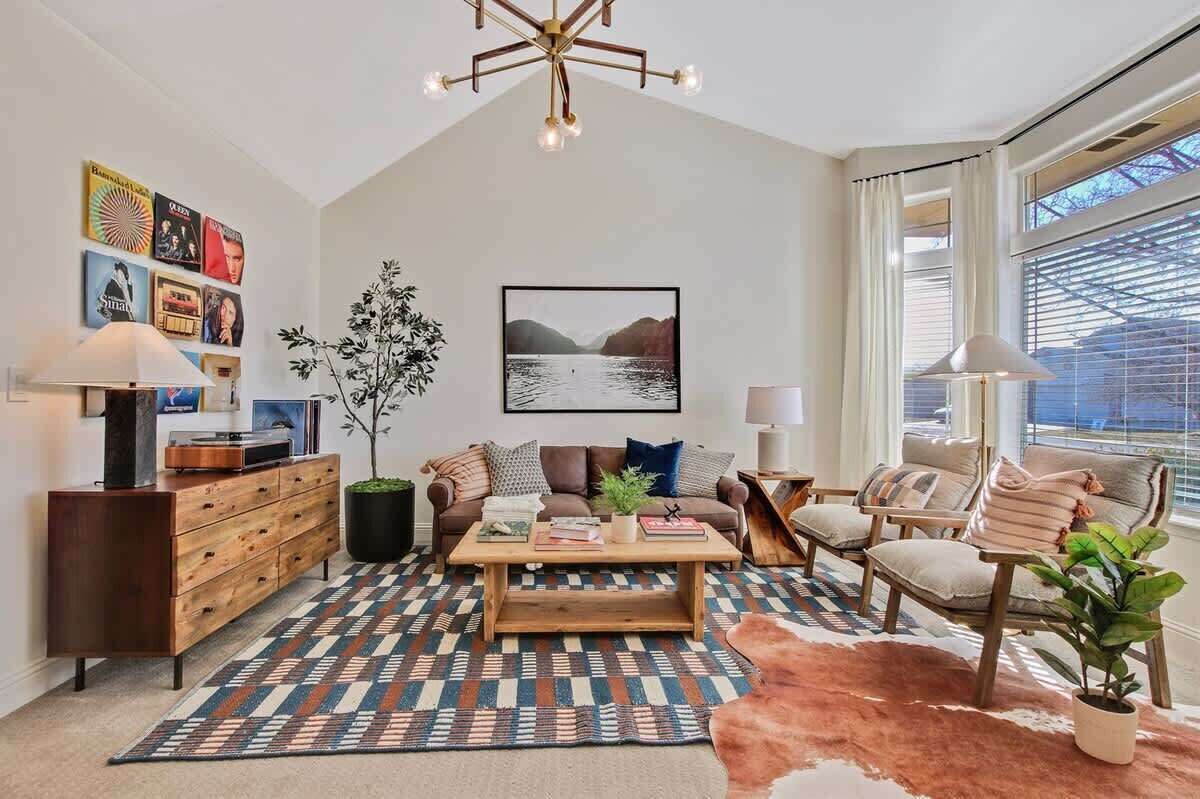 A modern living room with a wooden coffee table, a sofa, and a chair. There is a large window with curtains, a plant, and a wall-mounted picture.