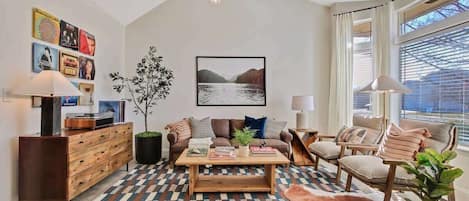 A modern living room with a wooden coffee table, a sofa, and a chair. There is a large window with curtains, a plant, and a wall-mounted picture.