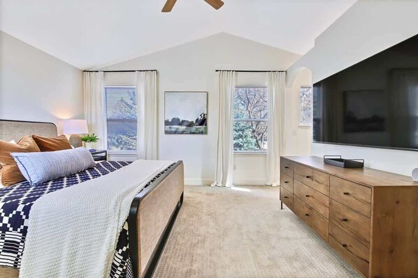 A bedroom with a bed, a dresser, a TV, and two windows with curtains.