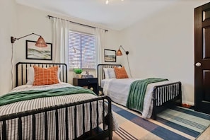 Un dormitorio con dos camas, cada una con ropa de cama y almohadas a rayas, una ventana con persianas, una mesita de noche con una lámpara y una puerta.
