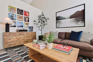 A living room with a brown sofa, a wooden coffee table with books and a plant, a wooden dresser, and a wall with framed pictures and a large landscape painting.