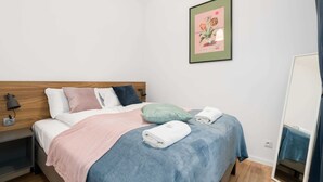 A neatly made bed with a blue and pink blanket, a green pillow, and a white towel. There is a framed picture on the wall and a mirror on the right.