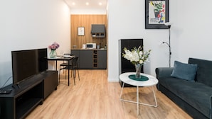 A modern living room with a black sofa, a small round table with a vase of flowers, a TV on a stand, and a kitchen area in the background.