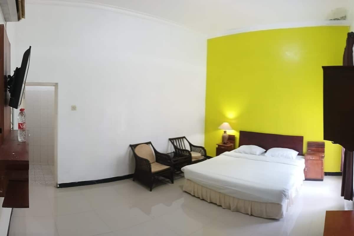 Quarto de hotel com cama, duas cadeiras, mesa de cabeceira com abajur e escrivaninha de madeira com uma garrafa.
