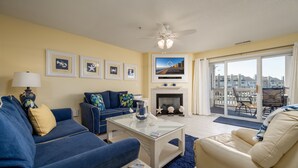 A living room with a blue sofa, a beige sofa, a coffee table, and a fireplace. There are framed pictures on the wall and a television above the fireplace. - Stunning Marsh and Bay Views from Beautiful 2 Bedroom Condo (Ocean City)
