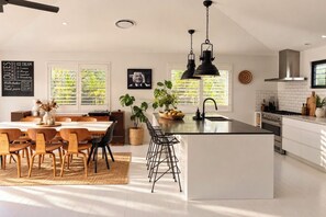 A modern kitchen with a central island, bar stools, a dining area with a long table, and a hanging light fixture.