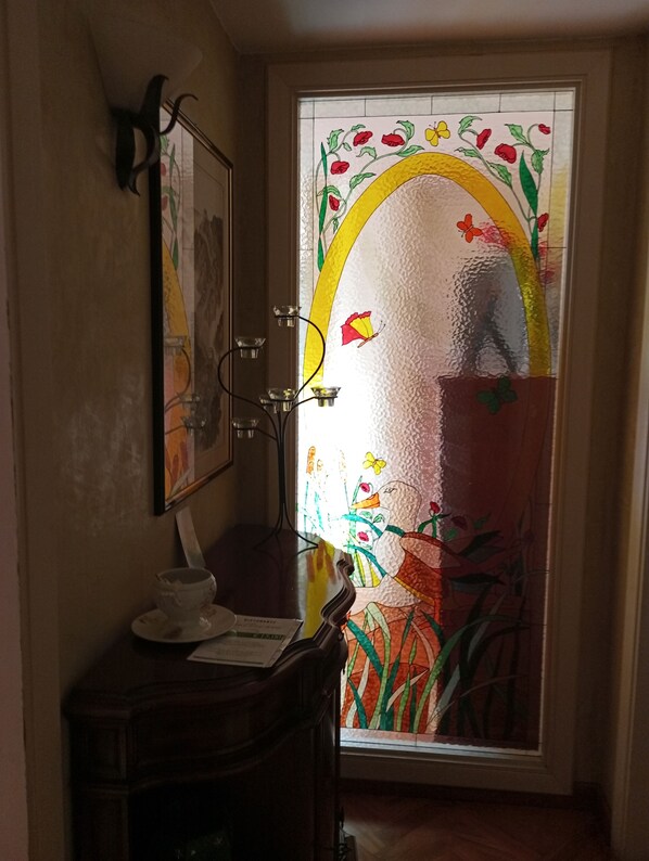 A room with a stained glass window featuring floral and bird designs, a wooden desk with a cup and papers, and a framed picture on the wall. - La Terrazza Apartment (Catania)