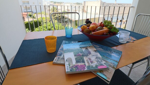 Un balcón con una mesa dispuesta para comer, que incluye un frutero, una copa y revistas.