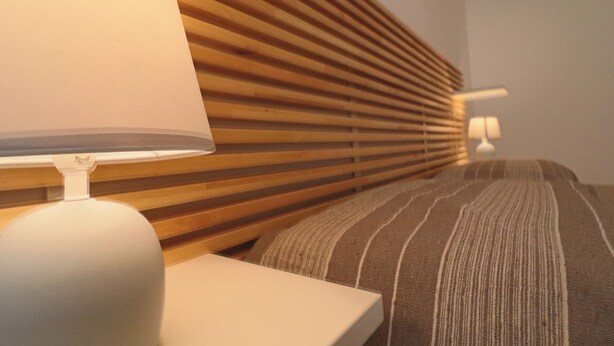 Una habitación de hotel ordenada con cabecera de madera, almohada a rayas y mesita de noche blanca con lámpara.
