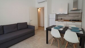 Un espacio habitable compacto con un sofá gris oscuro, una mesa de comedor con sillas blancas y una kitchenette con estufa y refrigerador.