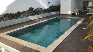 A rectangular swimming pool with a tiled deck, surrounded by a low white wall and a hedge.