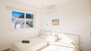A bedroom with two beds, a window with a view of buildings, a painting on the wall, and a ceiling fan. - Protaras holiday villa - rentprivatevillas 174 (Protaras)