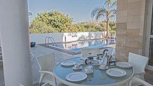 A glass-top table with white plates, cups, and a tea set, set against a backdrop of a swimming pool and palm trees. - Protaras holiday villa - rentprivatevillas 174 (Protaras)