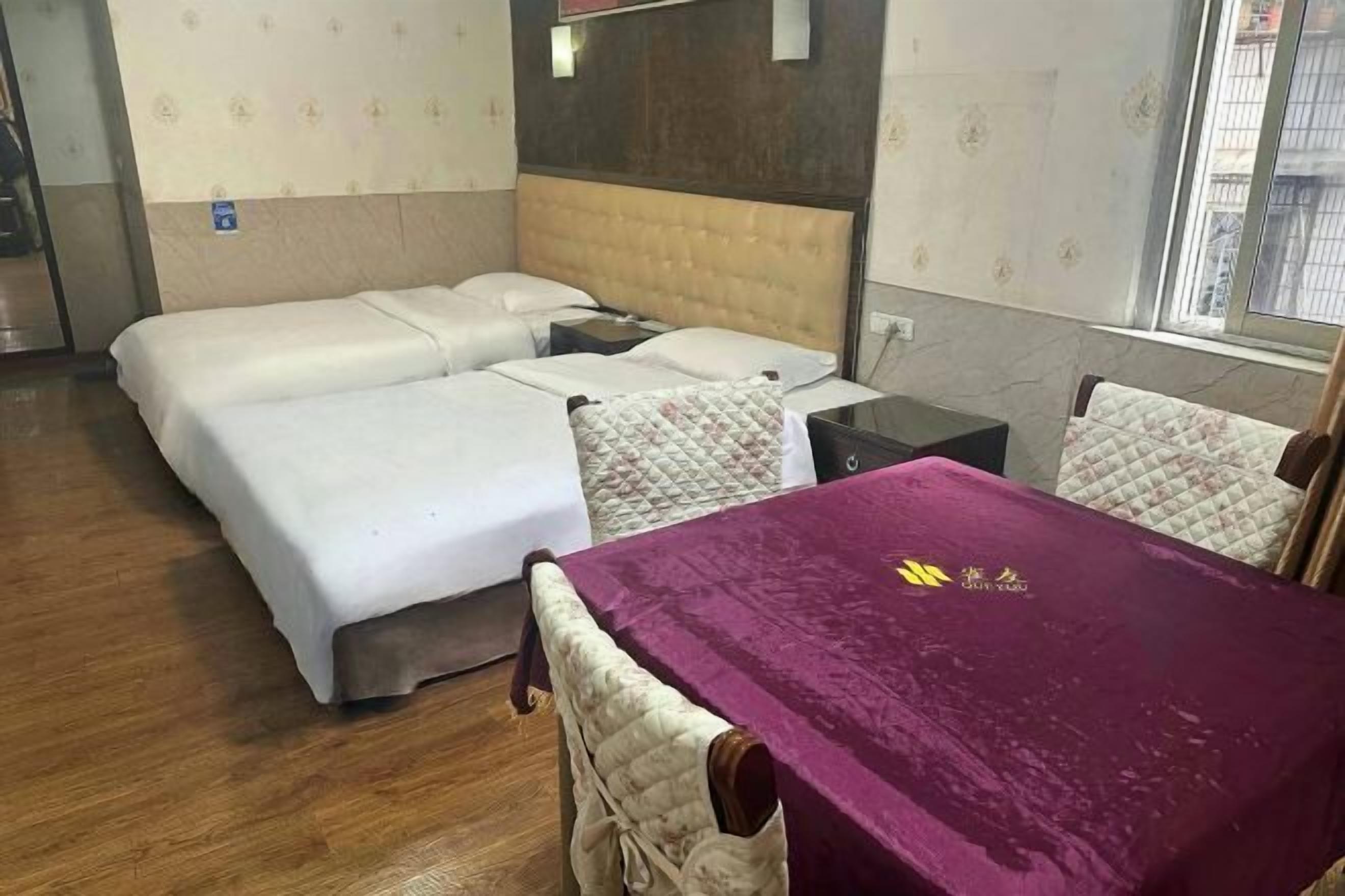 A hotel room with two beds, a small table with a purple tablecloth, and a window.