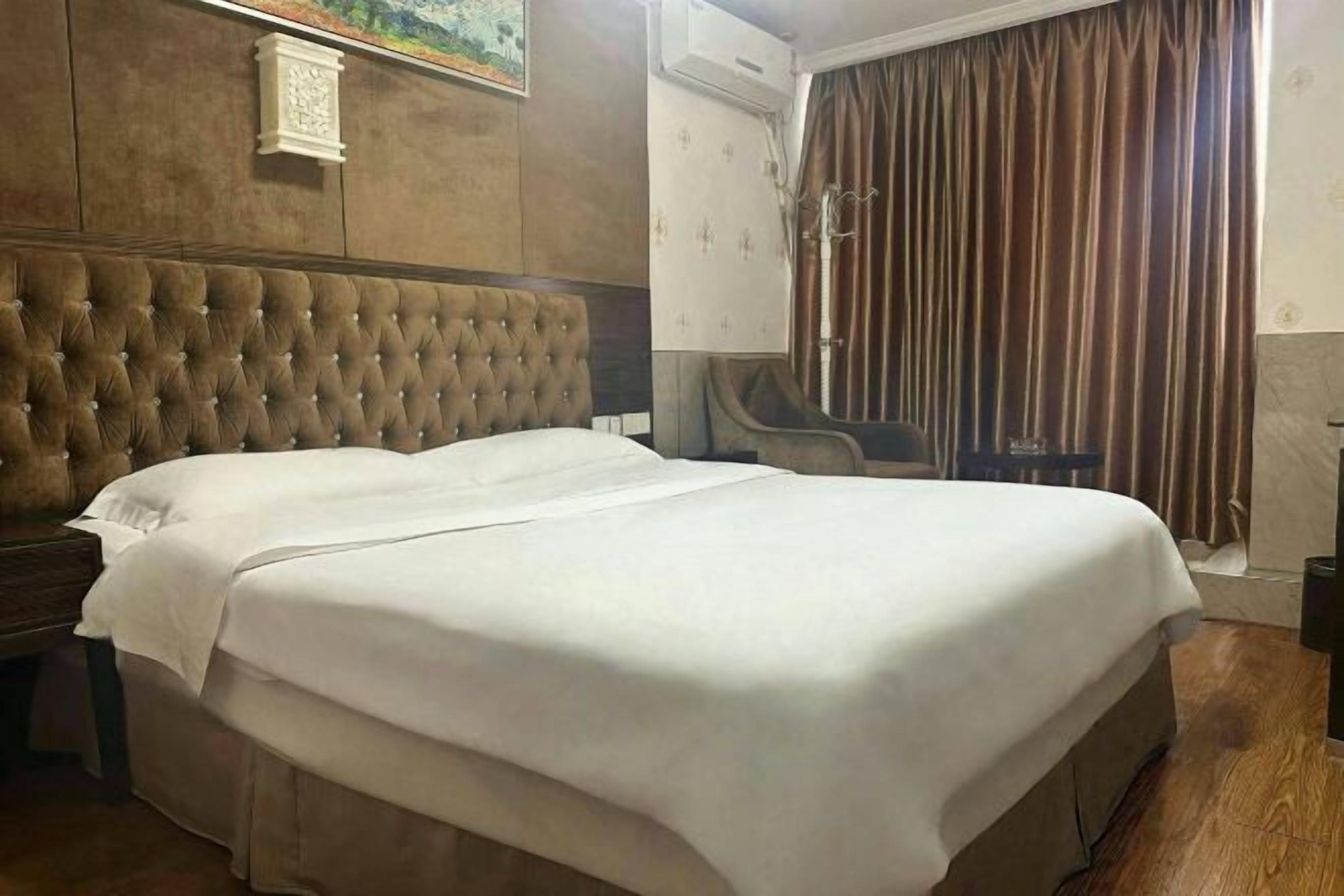 A double bed with a tufted headboard, a bedside table, a wall-mounted clock, and a window with curtains.