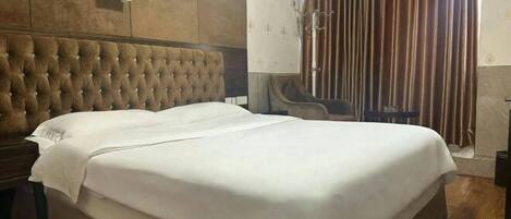 A double bed with a tufted headboard, a bedside table, a wall-mounted clock, and a window with curtains.