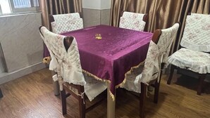 Un comedor con una mesa cuadrada cubierta con un mantel púrpura, rodeada por cuatro sillas tapizadas con tela blanca y detalles dorados.