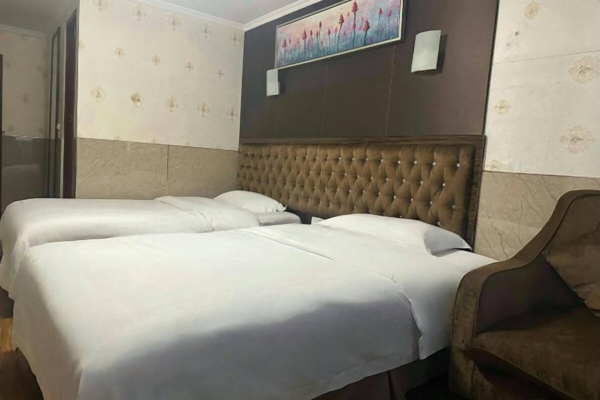 A hotel room with two beds, a large cushioned headboard, a painting on the wall, and a chair.