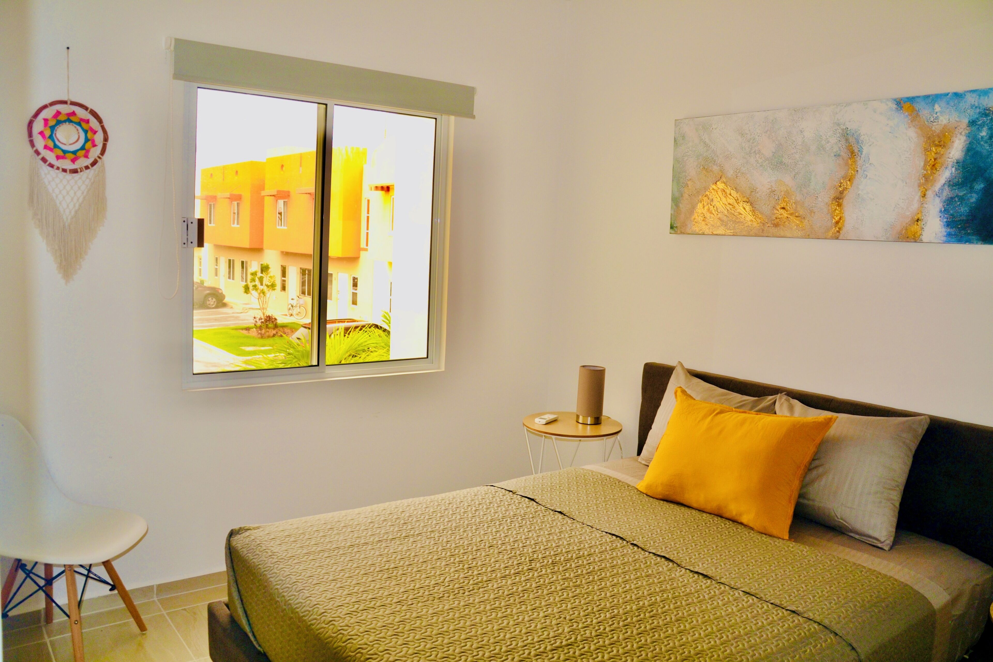 A hotel room with a bed, a small table, a chair, a window with a view of buildings, and a wall art piece.