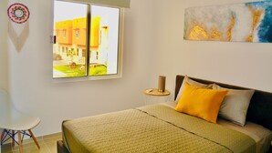 A hotel room with a bed, a small table, a chair, a window with a view of buildings, and a wall art piece.