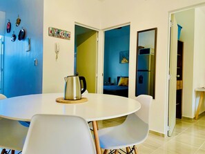 Uma sala de jantar com uma mesa redonda, cadeiras, chaleira e uma porta que dá para um quarto com cama e mesa de cabeceira.