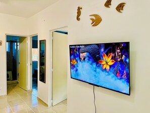 Um corredor com portas que dão para os quartos, uma televisão fixada na parede exibindo música Buda Flauta, e formas de crescente de madeira decorativas na parede.