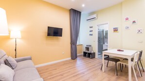 A living room with a sofa, a dining table with chairs, a TV mounted on the wall, and an air conditioning unit.