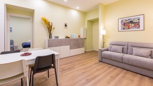 A modern living room with a grey sofa, a dining table with chairs, a kitchen area with a white cabinet, and a framed picture on the wall.