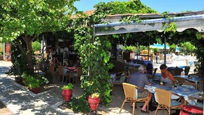 An outdoor café with tables and chairs under a shaded area, surrounded by greenery and a building in the background.