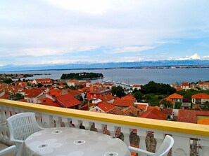 A balcony with white chairs and a table, overlooking a coastal town with red-roofed buildings and a marina. - Sea View Apartment Reno, Sleeps 5 (Preko)