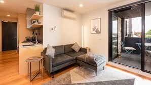 A modern living space with a grey sofa, a small kitchen area, and a sliding glass door leading to an outdoor patio. - Modern 1 Bedroom Apartment in Brunswick East Near CBD (Brunswick East)