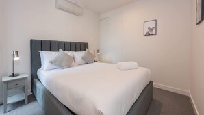 A neatly made bed with white bedding and gray pillows, a bedside table with a lamp, and a framed picture on the wall. - Modern 1 Bedroom Apartment in Brunswick East Near CBD (Brunswick East)