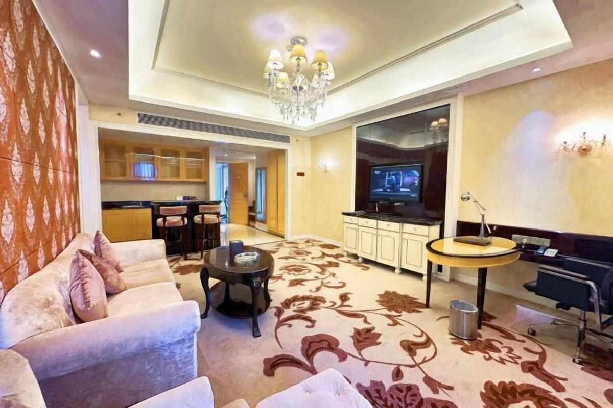 A hotel room with a sofa, a small round table, a TV, a kitchen area, and a chandelier.