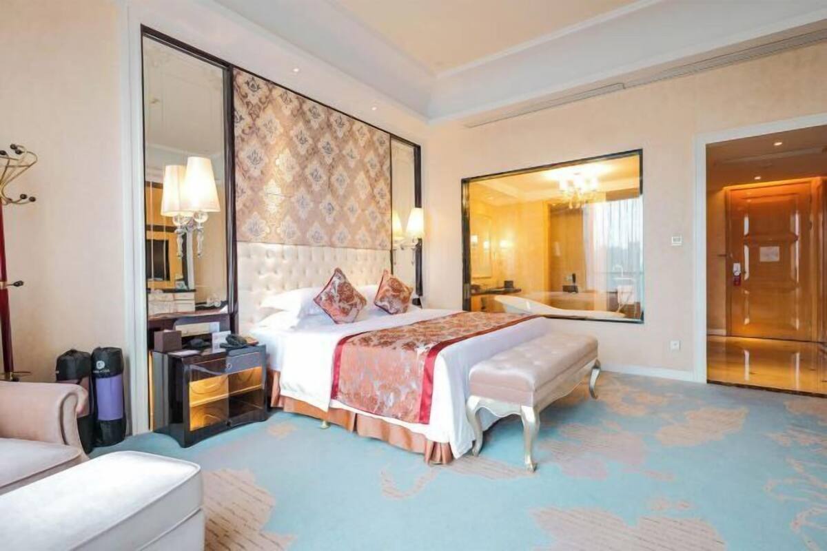 A hotel room with a large bed, a nightstand, a chair, a large mirror, and a bathroom visible through an open door.