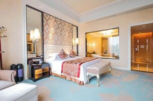 Quarto de hotel com cama grande, mesa de cabeceira, cadeira, espelho amplo e banheiro visível através de uma porta aberta.
