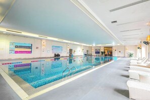 Una piscina interior con agua cristalina, rodeada de sillas de mimbre blancas y una zona de recepción con carteles informativos.