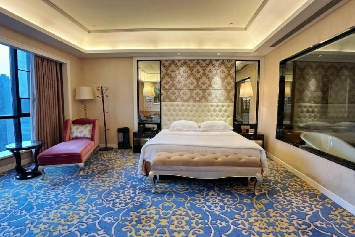 A hotel room with a large bed, a red chair, a floor lamp, a nightstand, and a large mirror.