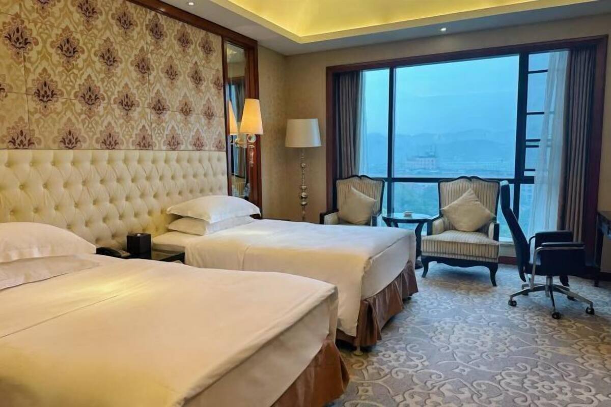 A hotel room with two beds, a large window with a view, a sitting area with a chair, and a patterned carpet.