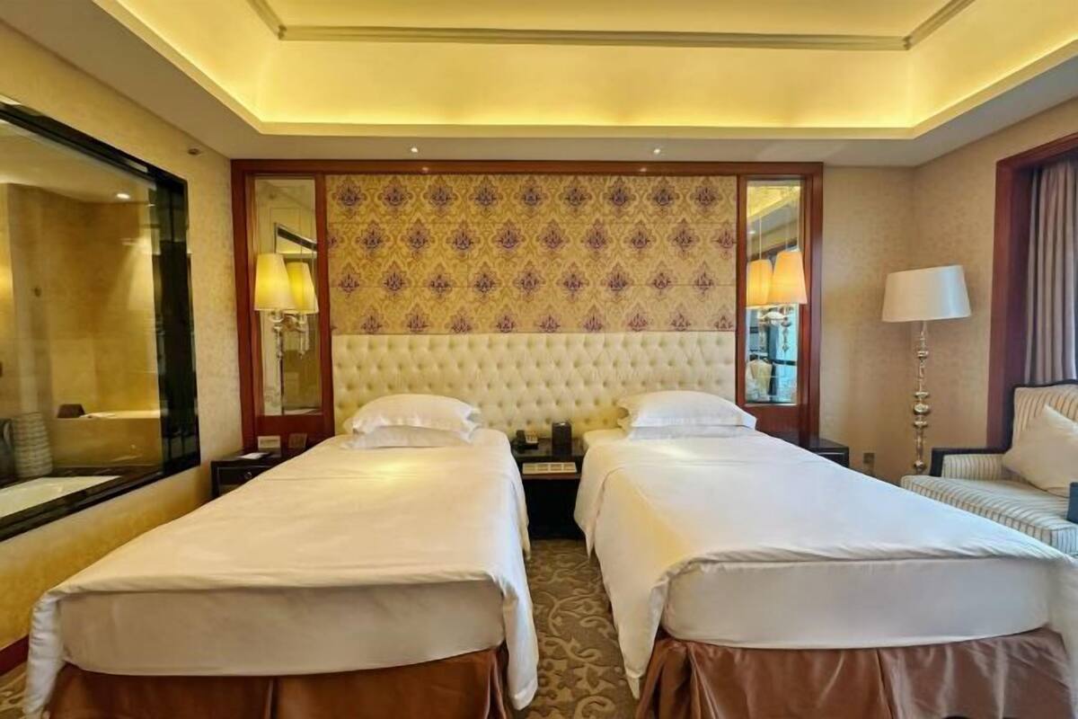 A hotel room with two single beds, a large mirror, a patterned headboard, and a small seating area with a lamp.