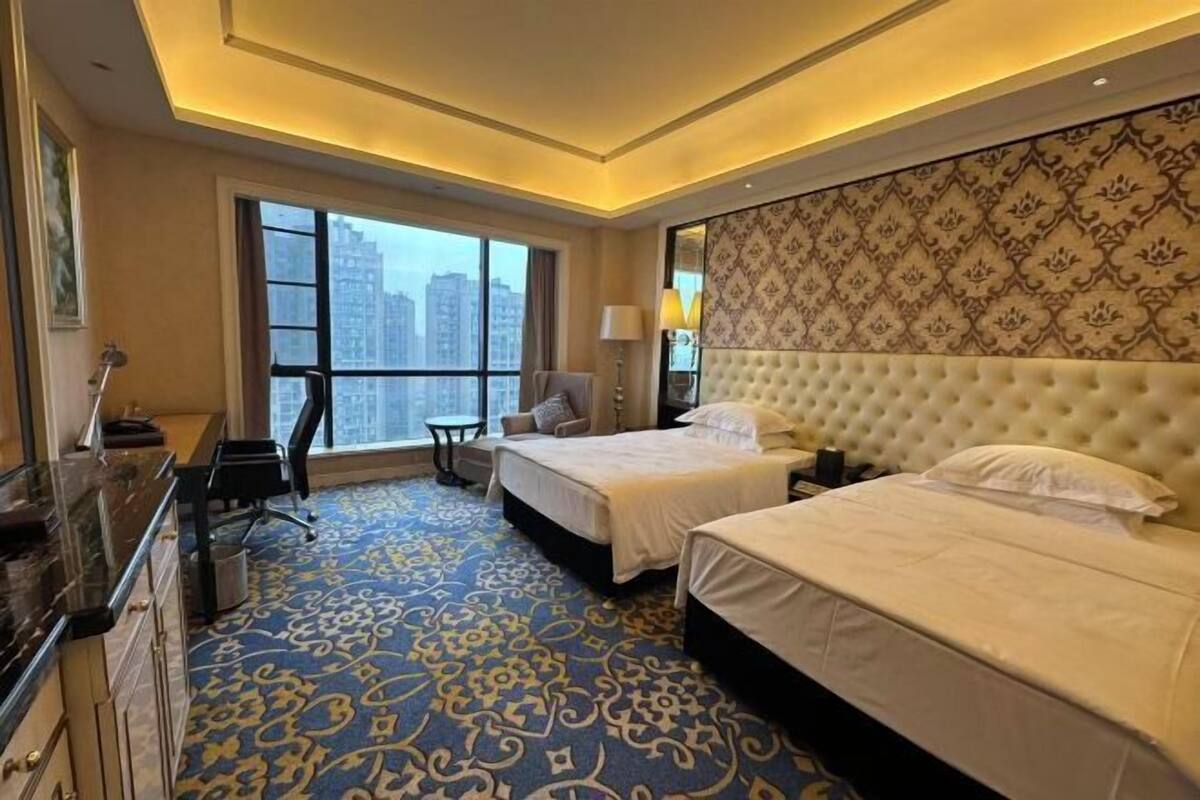 A hotel room with two beds, a desk, a chair, a large window, and a patterned wall.