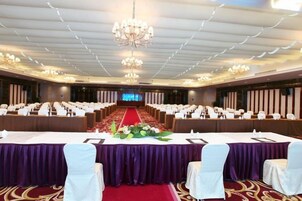 Un salón de conferencias con filas de sillas blancas, una larga mesa con mantel morado y una alfombra roja.