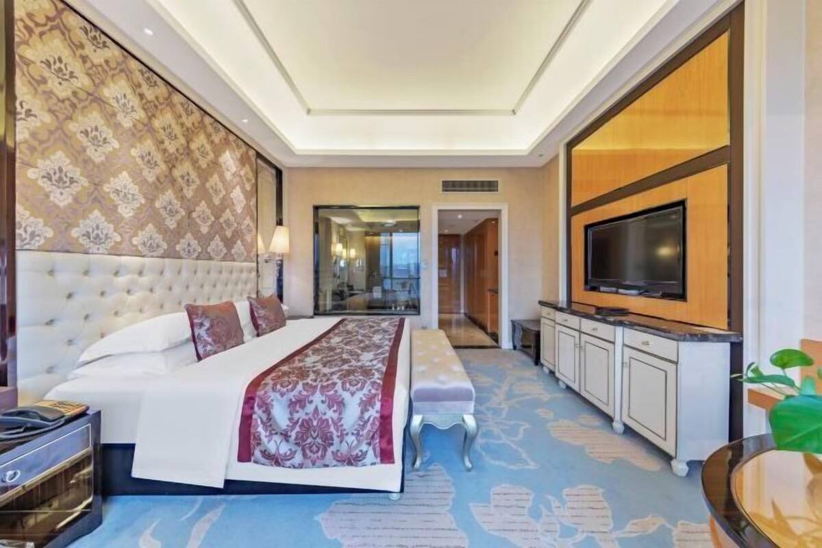 A hotel room with a large bed, a television, a minibar, and a bathroom visible through a glass door.