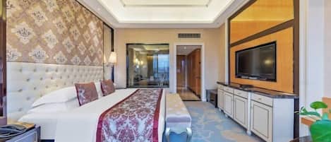 A hotel room with a large bed, a television, a minibar, and a bathroom visible through a glass door.