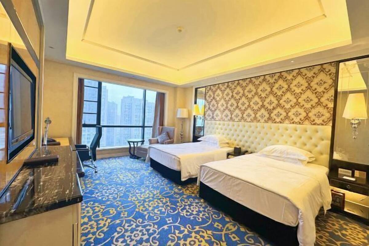 A hotel room with two beds, a desk, a chair, and a large window offering a city view.
